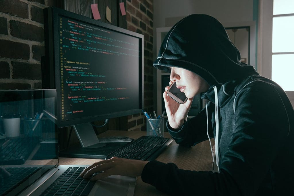 A person in a black hoodie eavesdropping on a phone conversation in front of a computer.