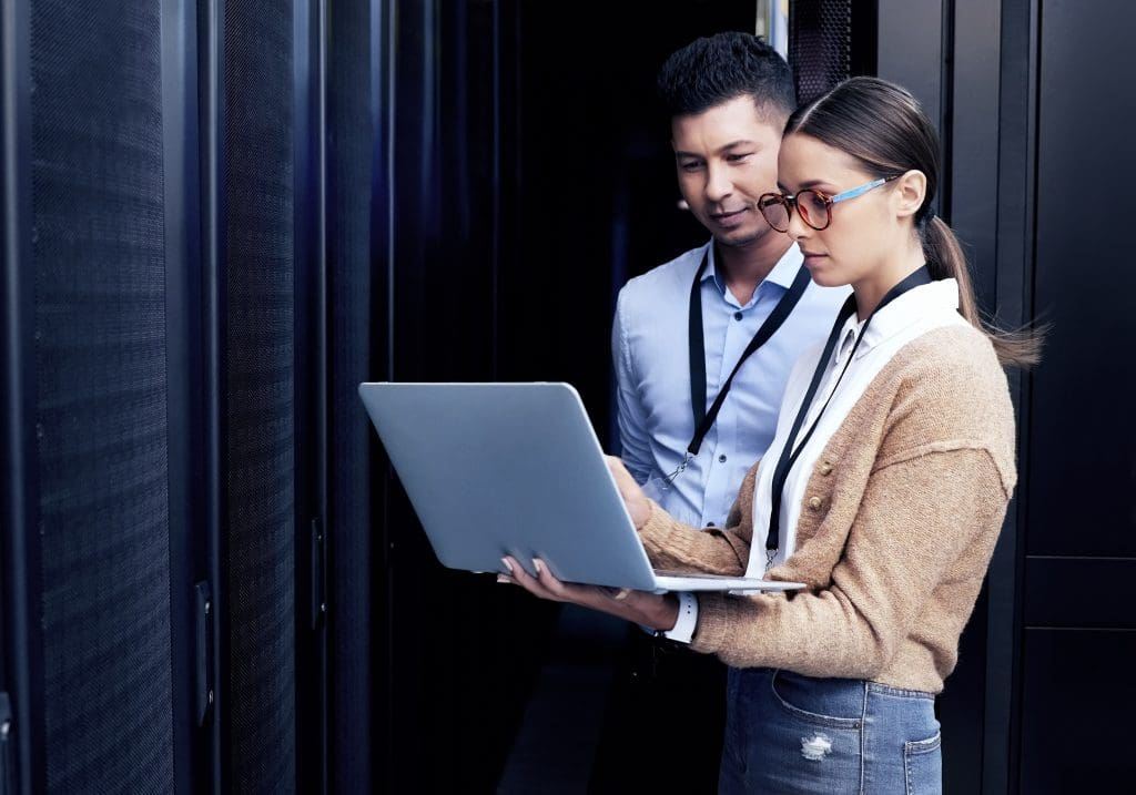 A man and woman in a server room examine a laptop in the woman's hands.